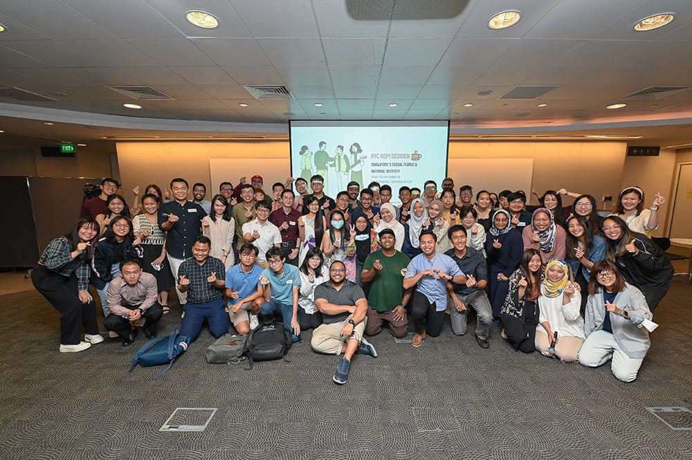 Large group posing in room with a "NYC Kopi Session" presentation screen visible.